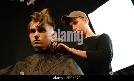 acconciatura e taglio di capelli da uomo con regolacapelli in un barbiere o in un parrucchiere. Servizio di parrucchiere in un moderno barbiere con illuminazione buia e vista dall'angolo basso con luce calda Foto Stock