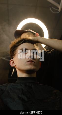 Il barbiere parrucchiere si occupa dell'acconciatura e dell'acconciatura con forbici e pettine. Concept Barbershop. angolo di visione basso in condizioni di luce buia con luce calda Foto Stock