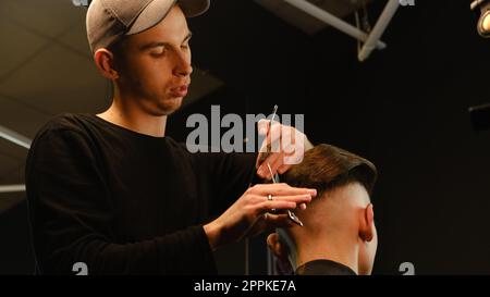 Il barbiere parrucchiere si occupa dell'acconciatura e dell'acconciatura con forbici e pettine. Concept Barbershop. vista posteriore con illuminazione a tasti scuri e luce calda Foto Stock