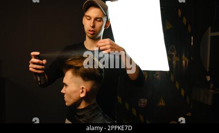 Il barbiere prepara lo styling dei capelli con lo spray dopo il taglio al barbiere. Giovane bell'uomo caucasico che ottiene un taglio di capelli in un moderno parrucchiere da parrucchiere professionale Foto Stock