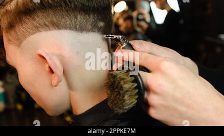 Scatto di un bel barbiere che fa un taglio di capelli al suo cliente usando il trimmer. Servizio di parrucchiere in un moderno barbiere con illuminazione buia e vista laterale calda e luminosa Foto Stock