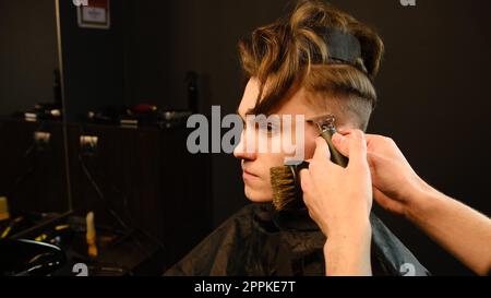 Scatto di un bel barbiere che fa un taglio di capelli al suo cliente usando il trimmer. Servizio di parrucchiere in un moderno barbiere con illuminazione buia e vista laterale calda e luminosa Foto Stock