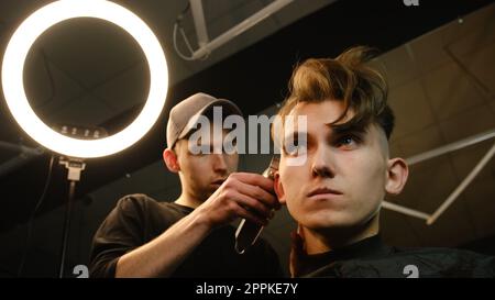 acconciatura e taglio di capelli da uomo con regolacapelli in un barbiere o in un parrucchiere. Servizio di parrucchiere in un moderno barbiere con illuminazione buia e vista dall'angolo basso con luce calda Foto Stock