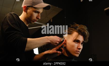 acconciatura e taglio di capelli da uomo con regolacapelli in un barbiere o in un parrucchiere. Servizio di parrucchiere in un moderno barbiere con illuminazione buia e vista laterale calda e luminosa Foto Stock