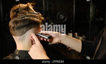 capelli da uomo e taglio con la macchina da barbiere, taglio per uomo. Servizio di parrucchiere con rasoio in un moderno barbiere con illuminazione buia e vista sulla schiena con luce calda Foto Stock