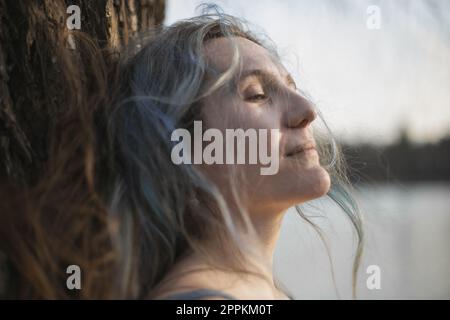 Primo piano grigio capelli donna con mezzo chiuso occhi appoggiati contro albero ritratto immagine Foto Stock