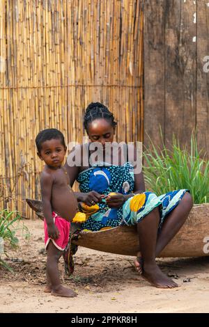 La donna malgascia che siede su una barca tradizionale dà frutta al suo bambino per la prima colazione. Foto Stock