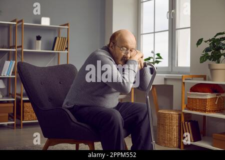 L'uomo anziano disabile esausto che si spiana appoggiandosi sulla canna mentre riposa sulla poltrona a casa. Foto Stock