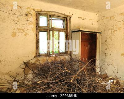 L'interno di un'antica casa rurale abbandonata con muri chiari incrinati, una vecchia bella finestra con vetri rotti e un pavimento coperto di sporcizia, rami e legno di pennello. Foto Stock