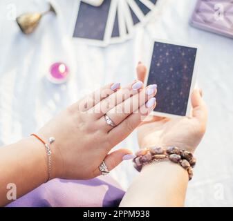 Donna tiene la mano con unghie viola su una carta Tarocchi su una superficie bianca con notebook e candela, vista dall'alto. Foto Stock