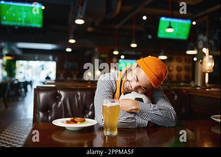 Divertente giovane uomo tifoso di calcio che dorme sulla palla mentre riposa nello sport bar Foto Stock