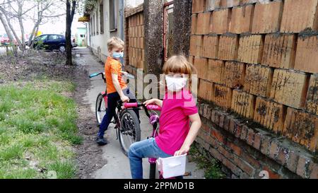 I bambini di 6 e 7 anni con maschere chirurgiche protettive bianche entrano per andare in bicicletta. Mettere in pausa il ciclo. Una ragazza con una maglietta rosa e un ragazzo biondo in arancione. Bambini per strada vicino alla vecchia recinzione in mattoni Foto Stock