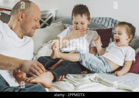 Il padre gioca con due bambini a letto. Papà fa il solletico ai piedi dei bambini. La famiglia di papà, ragazza e ragazzo Foto Stock