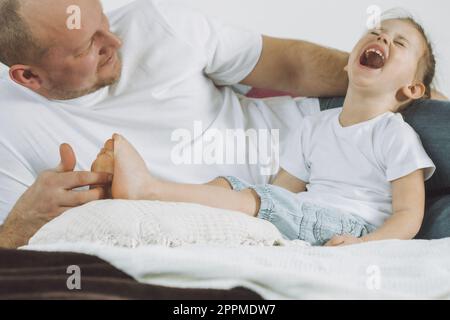 Il padre gioca con la figlia 2-4 sul letto. Papà fa il solletico ai piedi dei bambini. Famiglia, divertimento Foto Stock