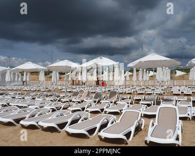Anapa, Russia, 15 agosto 2021 lettini e ombrelloni bagnati sulla spiaggia di mare durante una forte tempesta di pioggia. Ombrellone arrotolato sulla spiaggia contro il cielo nuvoloso scuro. Invasione di un tifone, ciclone o tempesta Foto Stock