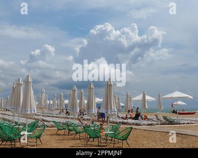 Anapa, Russia, 15 agosto 2021 sedie a sdraio e ombrelloni sulla spiaggia di sabbia dopo la pioggia. Stagione balneare. Caffè sulla spiaggia con sedie in vimini. Turisti che camminano. Un avvertimento tempesta. Nuvole di Cumulus Foto Stock