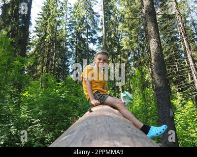 Un ragazzo biondo di 8 anni si siede sul tronco di un grande rotolo di albero di legno dolce caduto. Un bambino nel bosco. Il ragazzo si arrampicò sul tronco dell'albero. Avventure estive. Camminando nella foresta di abeti Foto Stock