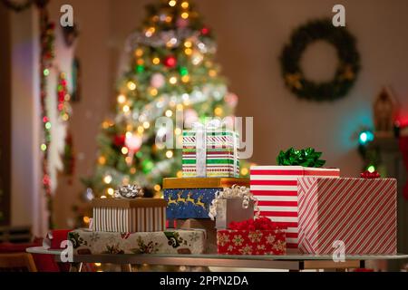 Regali di Natale disposti su un tavolo di fronte a un albero di Natale in una stanza della famiglia. Foto Stock