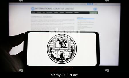 Persona che tiene il cellulare con il logo della Corte Internazionale di Giustizia (ICJ) sullo schermo di fronte alla pagina web. Messa a fuoco sul display del telefono. Foto Stock