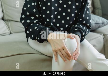 Primo piano giovane donna seduta sul divano e sensazione di dolore al ginocchio e sofferenza di dolore al ginocchio a casa. Concetto di assistenza sanitaria e dolore al ginocchio. Foto Stock