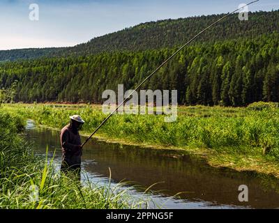 Un pescatore si trova nel fiume e cattura i pesci con una canna da pesca in legno. Pesca artigianale in Siberia. pesca con una canna dal tronco di una giovane tre Foto Stock