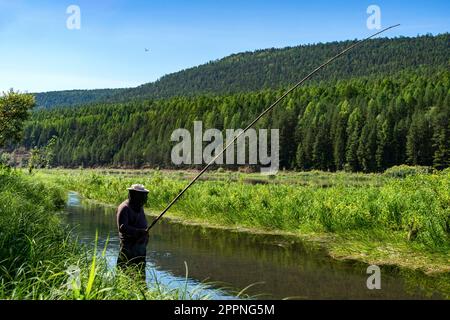 fisherman sorge nel fiume e cattura i pesci con la canna da pesca in legno. Pesca artigianale in Siberia. pesca con canna dal tronco di un albero giovane Foto Stock