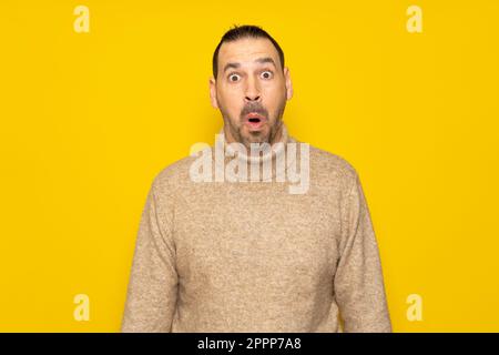 Barbuto ispanico uomo nel suo 40s indossando un dolcevita beige tremendamente stupito e sorpreso con bocca aperta in shock, isolato su sfondo giallo Foto Stock