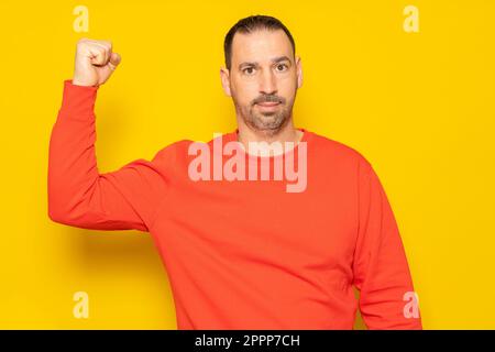 Uomo ispanico bearded che indossa una maglia rossa che flette il suo muscolo bicipite mostrando il suo stato pietoso di forma, isolato su sfondo giallo. Foto Stock