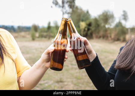 Primo piano di due amiche latine irriconoscibili che fanno un brindisi con le birre in campagna. Foto Stock