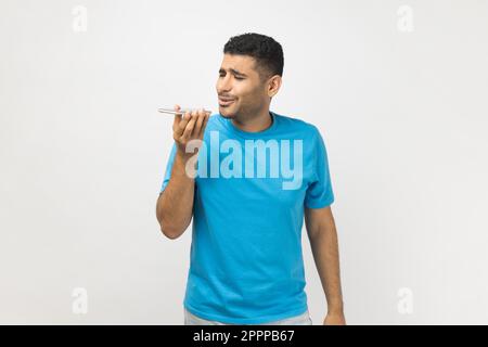 Ritratto di un bell'uomo non raso che indossa una T-shirt blu in piedi per tenere lo smartphone, registrare un messaggio vocale o utilizzare l'assistente vocale. Studio in interni isolato su sfondo grigio. Foto Stock