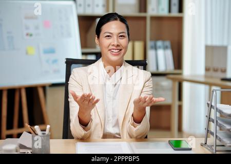 Giovane donna d'affari di successo in formalwear che spiega qualcosa a voi durante il seminario o la formazione mentre seduto sul posto di lavoro in ufficio Foto Stock