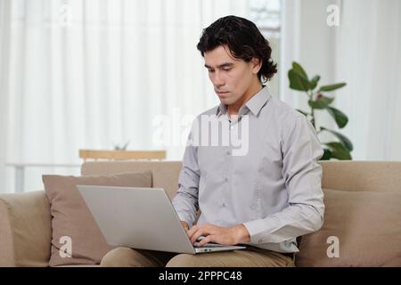 Analista giovane maschio che digita sulla tastiera del notebook mentre si siede sul divano in ufficio a casa e comunica con un collega online Foto Stock