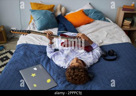 Riposante ragazzo afro-americano che suona la chitarra elettrica mentre si rilassa su un comodo letto matrimoniale dopo la scuola e si gode il tempo libero Foto Stock