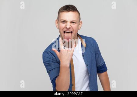 Ritratto di entusiasta ragazzo pazzo adolescente in camicia blu che mostra rock and roll gesto e uscire lingua, sensazione di essere fresco rocker in festival. Studio in interni isolato su sfondo grigio. Foto Stock