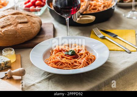 Tavolo con focalizzazione selettiva su un piatto di spaghetti con salsa di pomodoro. Pane, posate e bicchiere di vino rosso sullo sfondo Foto Stock