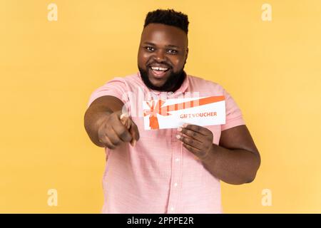 Ritratto di eccitato stupito uomo bearded indossando camicia rosa che tiene il vaucher regalo che punta alla macchina fotografica con il sorriso toothy, vincete il presente. Studio al coperto isolato su sfondo giallo. Foto Stock