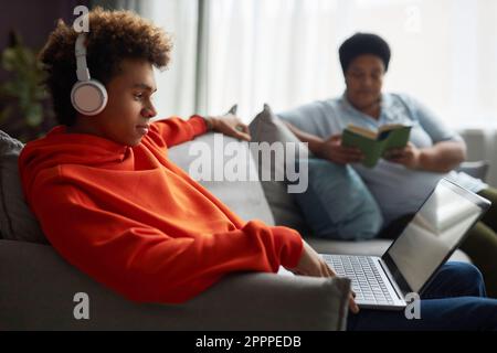 Vista laterale del ragazzo adolescente in cuffie e felpa con cappuccio rossa che guarda film o video online sullo schermo del notebook contro la donna che legge il libro Foto Stock