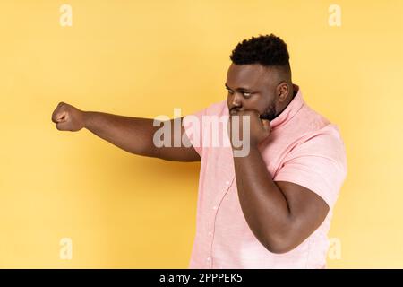 Ti colpisco. Vista laterale dell'uomo bearded che indossa una camicia rosa in piedi con pugilato in rilievo pronto a pugni, pronto a combattere, spirito di combattimento. Studio al coperto isolato su sfondo giallo Foto Stock