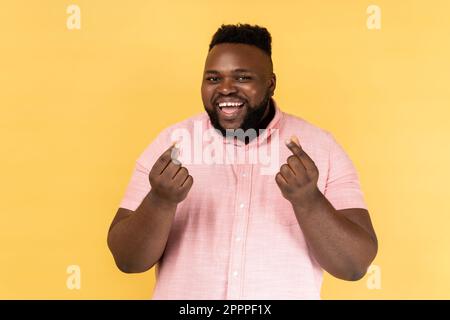 Ritratto di uomo sorridente bearded che indossa la camicia rosa fa il gesto dei soldi, sfrega le dita, guardando la macchina fotografica con espressione felice. Studio al coperto isolato su sfondo giallo. Foto Stock