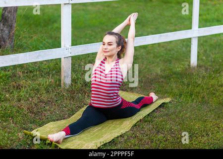 Ritratto completo dello yoga giovane atletico attraente donna che pratica lo yoga nel parco, facendo l'esercitazione del Dio della scimmia, le spaccature, la posa di Hanumanasana, esercitandosi fuori, portando lo sportswear. Foto Stock