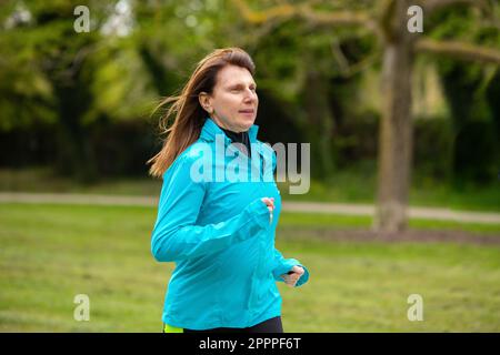 Runner femminile maturo che corre in un parco verde. Donna caucasica dai capelli rossi, in vestito sportivo, mantenersi in forma correndo in natura. Foto Stock
