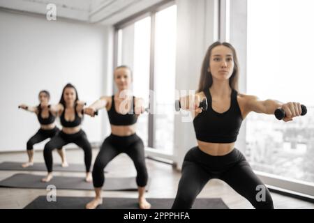 Giovani atleti in abbigliamento yoga con manubri mentre fanno squat su tappetini neri in palestra. Belle donne slim che eseguono esercizi di allenamento inferiore del corpo insieme in sala di allenamento. Foto Stock