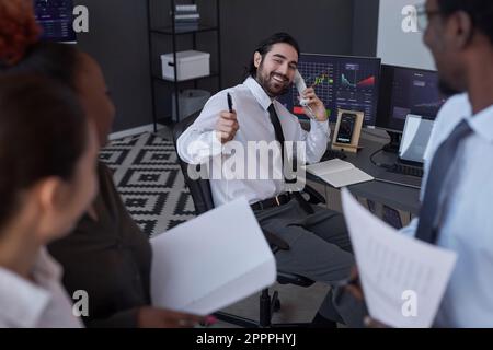 Giovane broker sorridente che vende le scorte con successo al cliente durante la conversazione al telefono Foto Stock