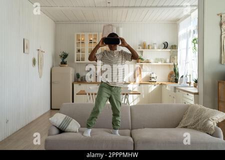 Il ragazzo afro-americano interessato gioca il video gioco si alza sul divano a casa in un casco per realtà virtuale. Foto Stock