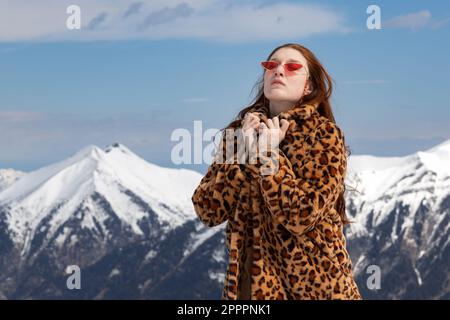 Un modello in un cappotto e occhiali luminosi si alza con gli occhi chiusi sullo sfondo delle montagne, l'Austria Foto Stock
