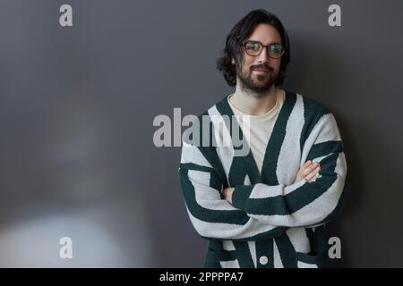 Ritratto di giovane uomo bearded in occhiali e maglione guardando la macchina fotografica in piedi su sfondo grigio Foto Stock