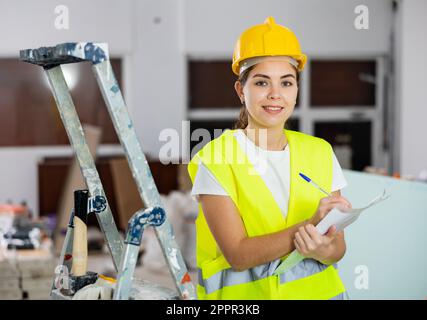 Ingegnere civile positivo femmina che prende appunti durante il controllo del cantiere Foto Stock