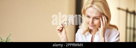 Donna d'affari matura stanca tolse i suoi occhiali, sentendo dolore, sovraccaricato di lavoro di computer in ufficio Foto Stock