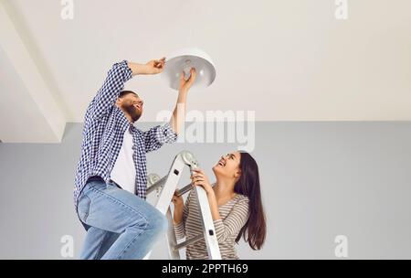 Felice coppia di famiglia che cambia una lampadina a risparmio energetico in una lampada da soffitto bianca a casa Foto Stock