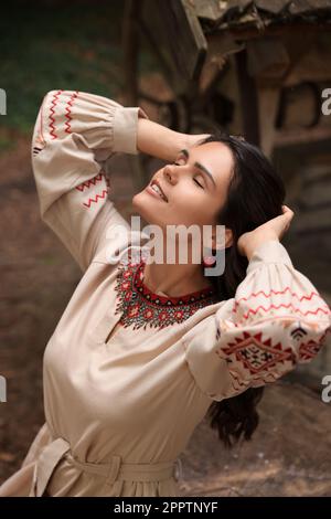 Bella donna con vestito ricamato e ornato collana con perline vicino al vecchio pozzo di legno in campagna. Vestiti nazionali ucraini Foto Stock